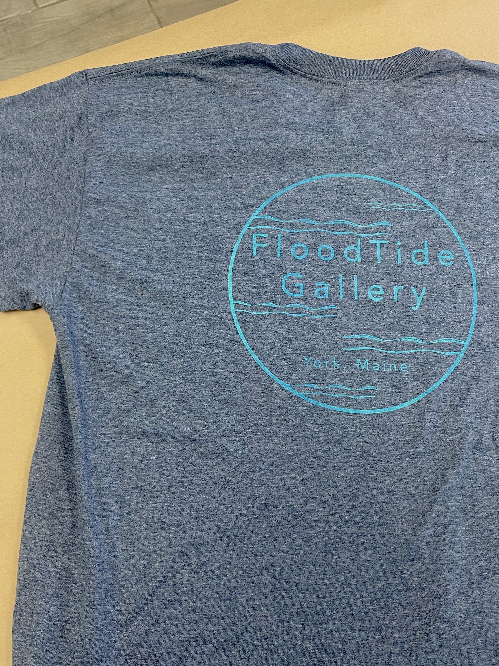 Blue t-shirt with 'Flood Tide Gallery' logo on a beige surface
